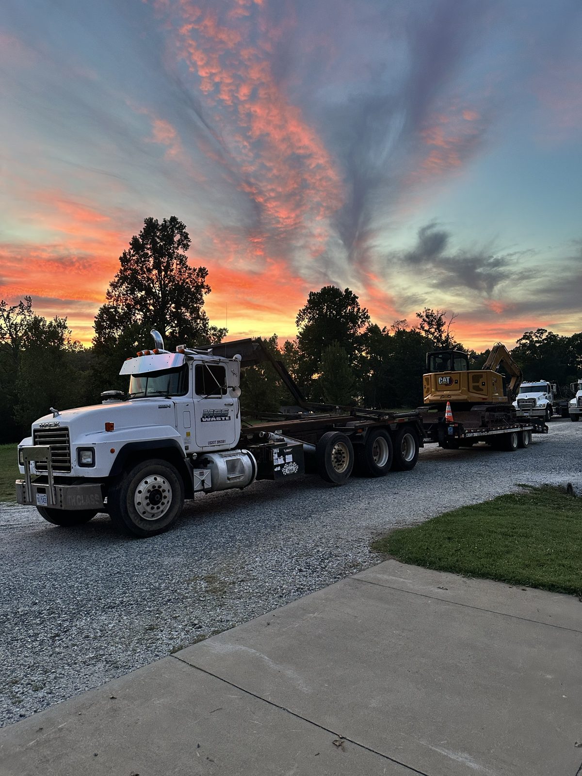 MGS Waste truck hauling an excavator on a lowboy trailer at sunset
